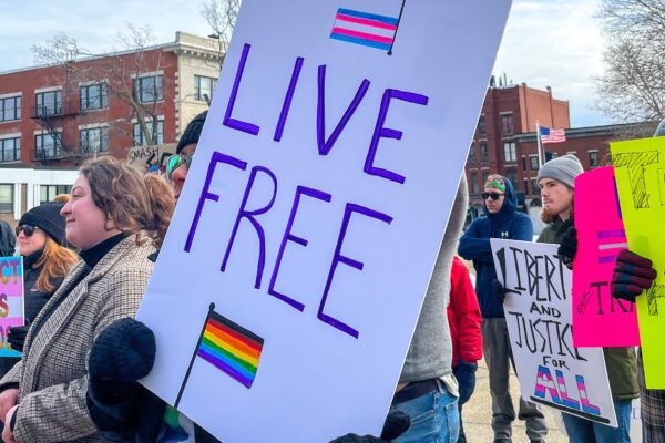 Protest sign reads Live Free and features Transgender flag and Rainbow flag