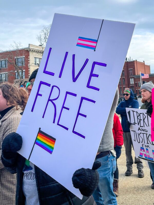 Protest sign reads Live Free and features Transgender flag and Rainbow flag