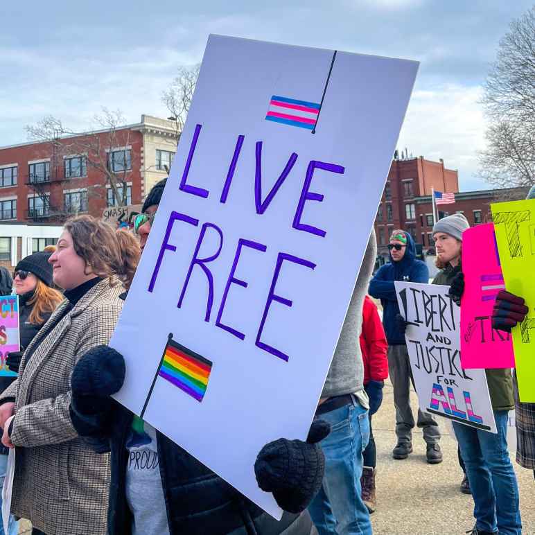 Protest sign reads Live Free and features Transgender flag and Rainbow flag 